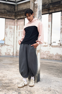 A model stands in a concrete warehouse wearing a dusty pink color super cropped hoodie with a black top, loose dark gray balloon pants, with sneakers.