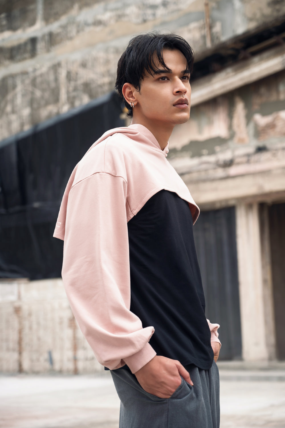 A man wearing a dusty pink colour super cropped hoodie with a black top underneathe, paired with gray pants, standing in front of a rustic stone building.